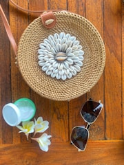 A compact straw crossbody bag hanging from a driftwood branch with seashells nearby.