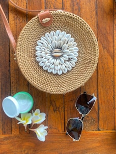 A compact straw crossbody bag hanging from a driftwood branch with seashells nearby.