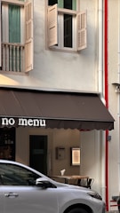An urban street scene featuring a building exterior with an awning displaying the words 'no menu.' Open windows with shutters are visible on the upper floor, and a parked white car is in the foreground. The building has a white facade with some architectural details, and a red pipe runs vertically along the wall.