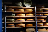 Multiple wheels of cheese are stacked on wooden shelves arranged in rows. The cheeses vary in size and have colorful labels with text and designs. The lighting creates a warm and rustic atmosphere, emphasizing the rich, creamy hues of the cheese.