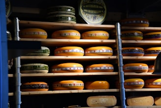 Multiple wheels of cheese are stacked on wooden shelves arranged in rows. The cheeses vary in size and have colorful labels with text and designs. The lighting creates a warm and rustic atmosphere, emphasizing the rich, creamy hues of the cheese.