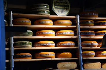 Multiple wheels of cheese are stacked on wooden shelves arranged in rows. The cheeses vary in size and have colorful labels with text and designs. The lighting creates a warm and rustic atmosphere, emphasizing the rich, creamy hues of the cheese.