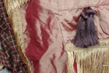 A close-up view of a fringed textile with intricate gold embroidery and braided tassels. The fabric is deep red, with hints of silky sheen, alongside a patterned fabric in shades of green and red.