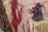A close-up view of a fringed textile with intricate gold embroidery and braided tassels. The fabric is deep red, with hints of silky sheen, alongside a patterned fabric in shades of green and red.
