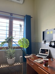 A cozy home office setup with a wooden desk featuring a laptop showing a video, a notebook with a pen, and a cup with a beverage. On the left, there is a wooden shelf holding two potted plants near a window with blue curtains. A pegboard with hooks and a pair of headphones is mounted on the wall above the desk. The room has an air conditioner unit visible on the wall.