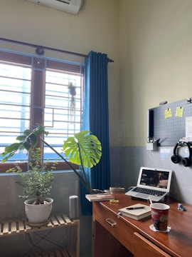 A cozy home office setup with a wooden desk featuring a laptop showing a video, a notebook with a pen, and a cup with a beverage. On the left, there is a wooden shelf holding two potted plants near a window with blue curtains. A pegboard with hooks and a pair of headphones is mounted on the wall above the desk. The room has an air conditioner unit visible on the wall.