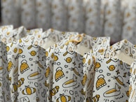 Gift bags featuring a playful pattern with cartoonish drawings, including cats, honeycomb patterns, hearts, bees, and hats. The design is predominantly yellow and black on a white background, with yellow strings for handles.