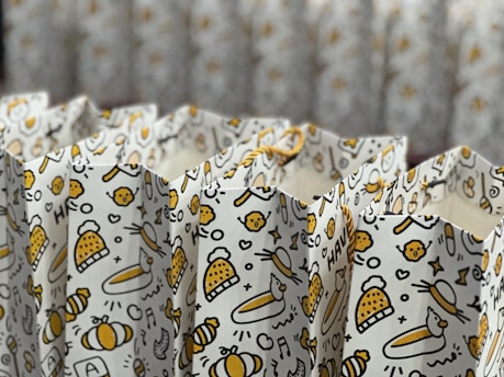 Gift bags featuring a playful pattern with cartoonish drawings, including cats, honeycomb patterns, hearts, bees, and hats. The design is predominantly yellow and black on a white background, with yellow strings for handles.