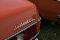 The rear end of a vintage car is featured with a close-up of the 'Automatic' emblem prominently displayed. The car is painted in a burnt orange color and the taillight assembly includes clear and red lenses. The setting appears to be an outdoor area with green grass visible.