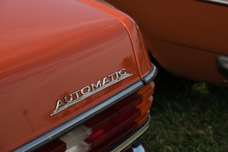 The rear end of a vintage car is featured with a close-up of the 'Automatic' emblem prominently displayed. The car is painted in a burnt orange color and the taillight assembly includes clear and red lenses. The setting appears to be an outdoor area with green grass visible.