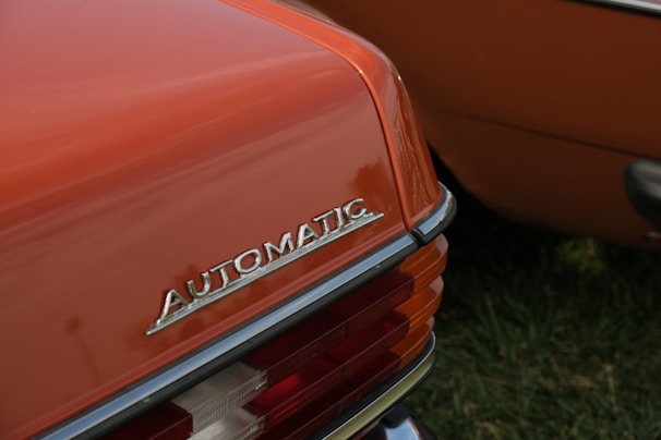 The rear end of a vintage car is featured with a close-up of the 'Automatic' emblem prominently displayed. The car is painted in a burnt orange color and the taillight assembly includes clear and red lenses. The setting appears to be an outdoor area with green grass visible.
