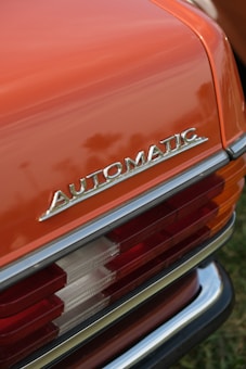 Close-up view of a vintage car's rear section with the word 'AUTOMATIC' prominently displayed in chrome lettering. The car features a shiny orange paint job and distinct tail lights with red and white sections. The bumper has a chrome finish.