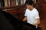 A person in a white t-shirt sits at a piano in a room lined with bookshelves filled with various books. The wooden furniture and shelves suggest a classic or vintage setting.