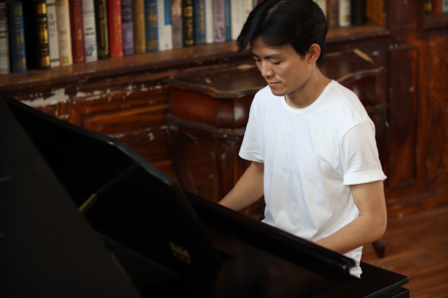 A person in a white t-shirt sits at a piano in a room lined with bookshelves filled with various books. The wooden furniture and shelves suggest a classic or vintage setting.