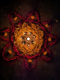 A circular pattern made with intricate designs featuring marigold petals and red powder, surrounding a central area with lit candles in decorative holders. The design is a traditional rangoli art piece set against a dark background.