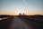 A straight path extends into the distance with a power plant visible on the horizon against the backdrop of a sunset. The foreground features a dirt road flanked by grassy fields and some scattered trees. Power lines stretch across the landscape, silhouetted against the colorful sky.
