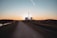 A straight path extends into the distance with a power plant visible on the horizon against the backdrop of a sunset. The foreground features a dirt road flanked by grassy fields and some scattered trees. Power lines stretch across the landscape, silhouetted against the colorful sky.