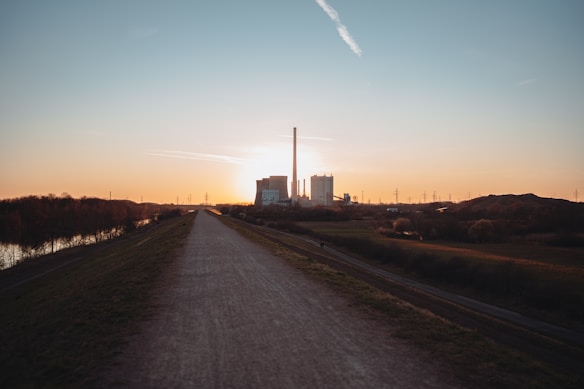 A straight path extends into the distance with a power plant visible on the horizon against the backdrop of a sunset. The foreground features a dirt road flanked by grassy fields and some scattered trees. Power lines stretch across the landscape, silhouetted against the colorful sky.