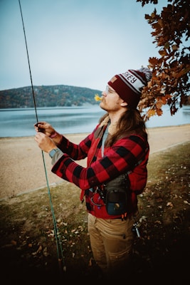 A person dressed warmly in a red and black plaid jacket is standing by a lake, holding a fishing rod. The individual is focused, appearing to adjust or prepare the fishing line. The background displays a calm body of water with a tree whose leaves are turning autumnal shades, suggesting a fall setting. The sky is overcast, adding to the serene atmosphere.
