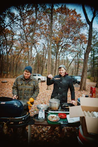 A candid photo of a smiling traveler cooking outdoors with a happy dog nearby, surrounded by nature.