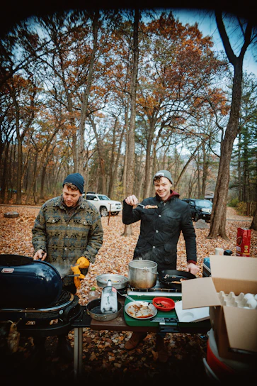 A candid photo of a smiling traveler cooking outdoors with a happy dog nearby, surrounded by nature.