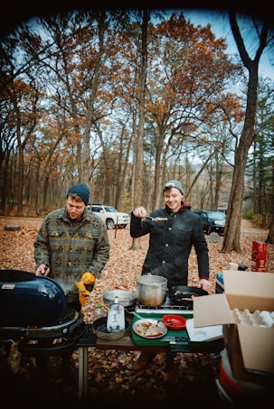 Two people are cooking outdoors in a forest setting. They are surrounded by trees with autumn foliage and are using a portable grill and various cookware on a table. There are eggs, a pot, and other cooking essentials visible. One person is smiling and holding a piece of food with a utensil, while the other is tending to the grill. Cars can be seen parked in the background.