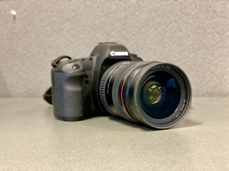 A DSLR camera with a large lens is positioned on a flat surface. The camera appears to be professional-grade with a prominent rubberized grip and various controls and dials on the body. The lens is a Canon EF model, as indicated by the branding on its rim.