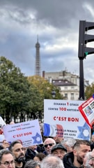 A group of people gathered for a protest or rally, holding signs with text in French. The background includes trees and partially visible historic buildings, with the Eiffel Tower prominently visible under a cloudy sky.