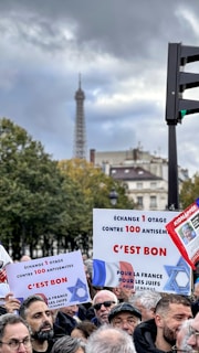 A group of people gathered for a protest or rally, holding signs with text in French. The background includes trees and partially visible historic buildings, with the Eiffel Tower prominently visible under a cloudy sky.