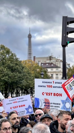 A group of people gathered for a protest or rally, holding signs with text in French. The background includes trees and partially visible historic buildings, with the Eiffel Tower prominently visible under a cloudy sky.
