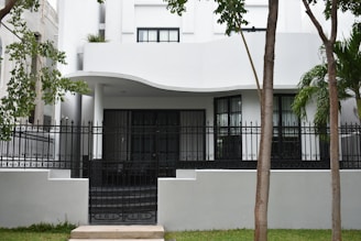 A modern building with a curved white facade and black-framed windows. The entrance is gated with ornamental black ironwork. Tall trees with green leaves are in the foreground, and there is a well-maintained lawn.