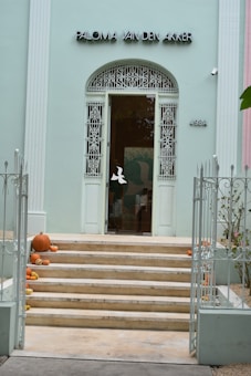 A pastel-colored building facade features an ornate wrought iron gate and a set of stone steps leading to a glass door. Pumpkins are arranged on the steps, adding a decorative touch.