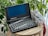 A vintage Gateway 2000 Handbook laptop sits on a wooden round table, with a lush green plant in the background. The screen is blank, and the device has an old-style keyboard. Next to the laptop, there's a transparent case filled with electronic components and a coiled red wire.