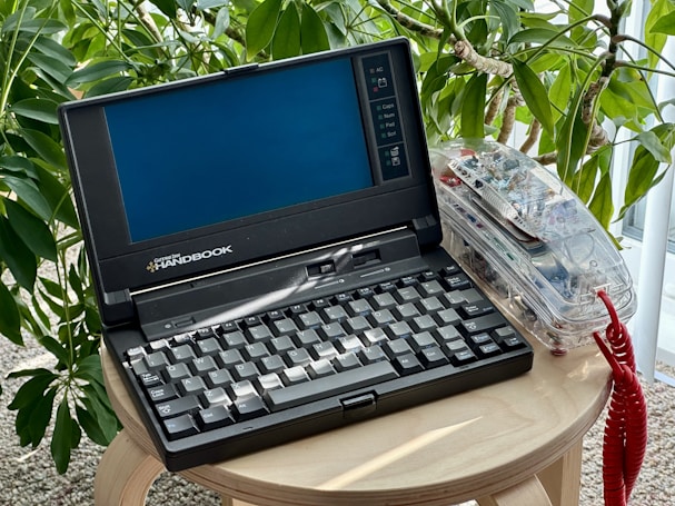 A vintage Gateway 2000 Handbook laptop sits on a wooden round table, with a lush green plant in the background. The screen is blank, and the device has an old-style keyboard. Next to the laptop, there's a transparent case filled with electronic components and a coiled red wire.