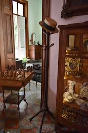 A vintage interior space featuring a wooden coat rack with a straw hat on it, a table with carved chess pieces, and a wooden cabinet displaying china dishes and antique items. The floor is adorned with ornate patterned tiles, and the setting is softly lit by natural light coming through a partially open window.