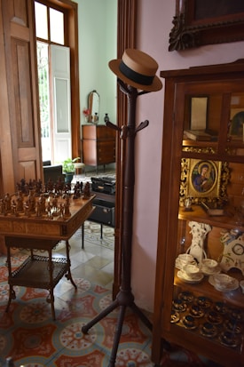 A vintage interior space featuring a wooden coat rack with a straw hat on it, a table with carved chess pieces, and a wooden cabinet displaying china dishes and antique items. The floor is adorned with ornate patterned tiles, and the setting is softly lit by natural light coming through a partially open window.