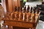 An intricately carved wooden chess set is displayed on a matching table with detailed engravings on its sides. The chess pieces are styled in elaborate medieval attire, suggestive of historical or mythical figures. The board is set for a game in natural light, with a hint of greenery and furniture in the background, adding to a cozy, antique setting.