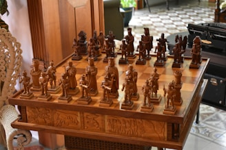 An intricately carved wooden chess set is displayed on a matching table with detailed engravings on its sides. The chess pieces are styled in elaborate medieval attire, suggestive of historical or mythical figures. The board is set for a game in natural light, with a hint of greenery and furniture in the background, adding to a cozy, antique setting.