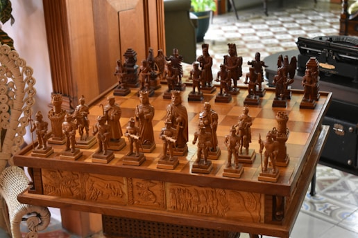 An intricately carved wooden chess set is displayed on a matching table with detailed engravings on its sides. The chess pieces are styled in elaborate medieval attire, suggestive of historical or mythical figures. The board is set for a game in natural light, with a hint of greenery and furniture in the background, adding to a cozy, antique setting.