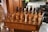 An intricately carved wooden chess set is displayed on a matching table with detailed engravings on its sides. The chess pieces are styled in elaborate medieval attire, suggestive of historical or mythical figures. The board is set for a game in natural light, with a hint of greenery and furniture in the background, adding to a cozy, antique setting.
