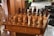 An intricately carved wooden chess set is displayed on a matching table with detailed engravings on its sides. The chess pieces are styled in elaborate medieval attire, suggestive of historical or mythical figures. The board is set for a game in natural light, with a hint of greenery and furniture in the background, adding to a cozy, antique setting.
