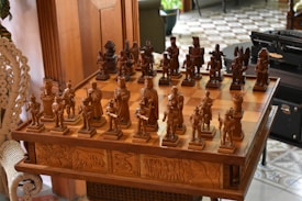 An intricately carved wooden chess set is displayed on a matching table with detailed engravings on its sides. The chess pieces are styled in elaborate medieval attire, suggestive of historical or mythical figures. The board is set for a game in natural light, with a hint of greenery and furniture in the background, adding to a cozy, antique setting.