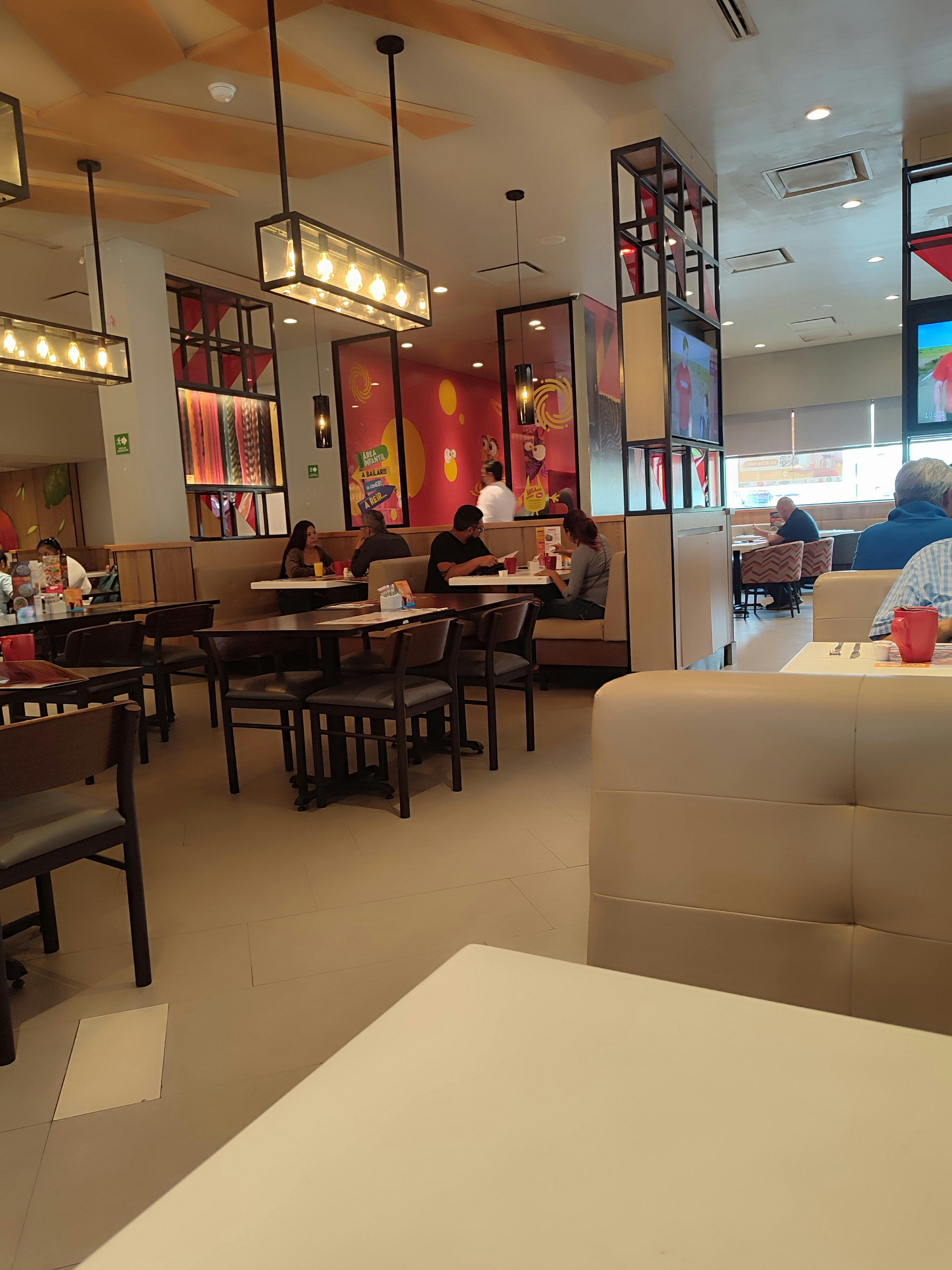 A modern restaurant interior featuring wooden furniture, pendant lighting, and colorful murals on the walls. There are several patrons seated at different tables, creating a casual dining atmosphere.