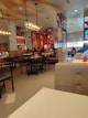 A modern restaurant interior featuring wooden furniture, pendant lighting, and colorful murals on the walls. There are several patrons seated at different tables, creating a casual dining atmosphere.