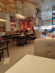 A modern restaurant interior featuring wooden furniture, pendant lighting, and colorful murals on the walls. There are several patrons seated at different tables, creating a casual dining atmosphere.