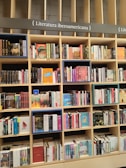 A bookshelf filled with a variety of books categorized under Ibero-American literature. The shelves are organized with books displayed upright and stacked, showcasing colorful covers and titles. The structure is wooden with sections divided by horizontal and vertical beams.