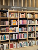 A bookshelf filled with a variety of books categorized under Ibero-American literature. The shelves are organized with books displayed upright and stacked, showcasing colorful covers and titles. The structure is wooden with sections divided by horizontal and vertical beams.