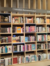 A bookshelf filled with a variety of books categorized under Ibero-American literature. The shelves are organized with books displayed upright and stacked, showcasing colorful covers and titles. The structure is wooden with sections divided by horizontal and vertical beams.