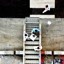 Aerial view of three people in uniforms on a staircase, with one person carrying a tray. The scene takes place outdoors with a concrete surface and painted markings visible around the stairs.