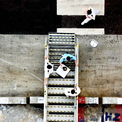 Aerial view of three people in uniforms on a staircase, with one person carrying a tray. The scene takes place outdoors with a concrete surface and painted markings visible around the stairs.